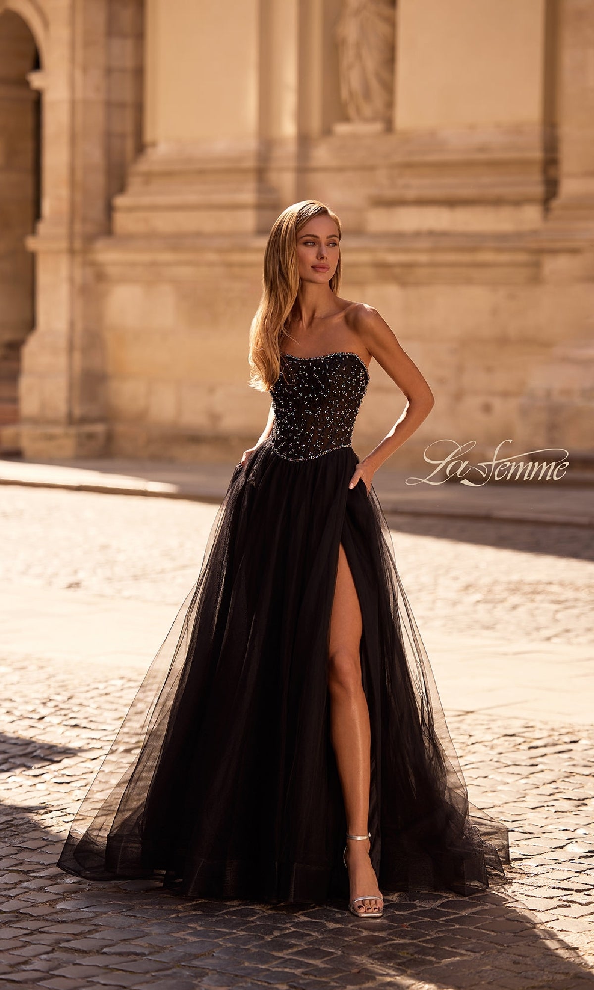 A woman in the La Femme 32856 Strapless Long Black Prom Ball Gown with a beaded bodice and high slit stands outdoors on a cobblestone street in front of a stone building.